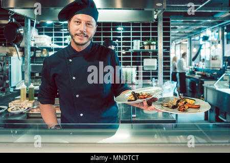 Ritratto di un fiducioso maestro chef tenendo due piastre con il cibo gustoso Foto Stock