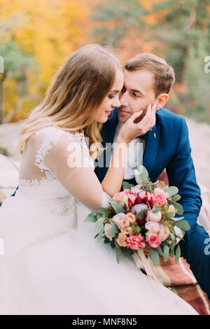 Vista ravvicinata della sposa accarezzare la guancia dello sposo mentre è seduto sulla roccia. Foto Stock