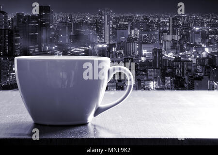 Caffè caldo in tazza bianca sul contatore di legno bar accanto al vetro della finestra su alta torre con osaka city night view. Effetto monotona. Foto Stock