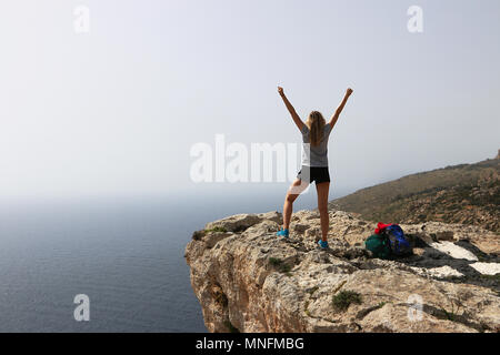 Trionfa il giovane donna a braccia alzate celebra dopo una salita con successo. In piedi torna alla telecamera che guarda al mare dal ciglio della scogliera. Copia dello spazio. Foto Stock