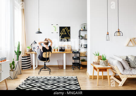 Giovane donna freelance che lavorano in remoto a designer home in accogliente ufficio bianco camera con lampadari, muro bianco, finestra grande, confortevole lettino e aloe pla Foto Stock