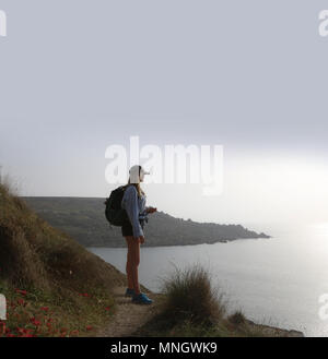 Giovane donna in piedi che guarda al mare, indossa pantaloncini uno zaino e tappo di picco. Ella è in alto guardando verso il basso Foto Stock