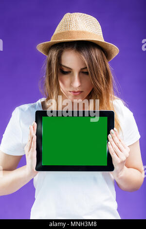 Sorridente ragazza bionda vestita di bianco t-shirt e hat detiene il tablet PC con schermo verde prima di sfondo blu, pubblicizzare il concetto di spazio Foto Stock