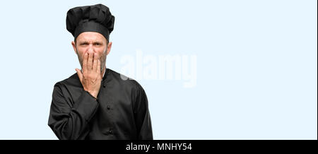 Senior uomo cook, indossando chef hat copre bocca in stato di shock, guarda timido, esprimendo il silenzio e confondere i concetti, spaventata isolate su sfondo blu Foto Stock