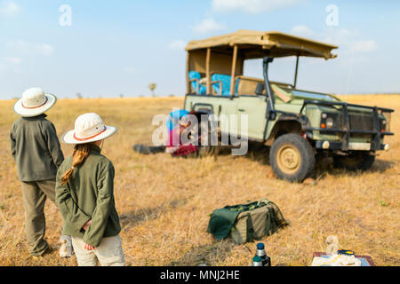 I bambini in Kenya safari al mattino game drive in veicolo aperto guardando guide masai cambiando il pneumatico sgonfio Foto Stock