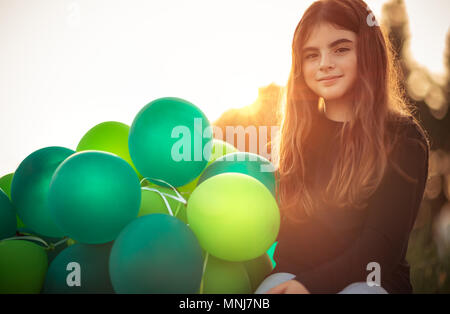 Ritratto di una incantevole piccola ragazza seduta nel parco con la pila di verde palloncini aria all'aperto in serata dolce della luce del sole, festeggia il compleanno, preparati Foto Stock