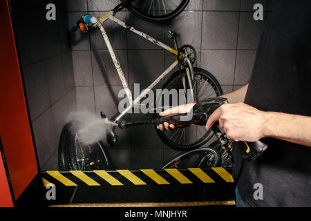 Servizi Lavaggio professionale di bicicletta in officina. Close-up di mano giovani caucasici uomo elegante bicicletta facendo pulizia utilizzando un sistema automatico di pompa elettrica dell'acqua. Spray scatter dalla pressione Foto Stock