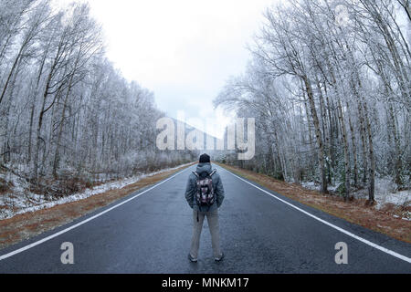 L'uomo in grigio con uno zaino in piedi al centro della strada grigio in una foresta innevata. Foto Stock