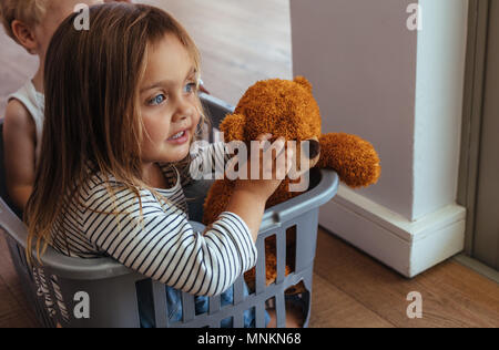 Bella ragazza seduta in un cestello di lavaggio con il suo orsacchiotto e fratello all'indietro. I fratelli giocando in un cestello di lavaggio a casa. Foto Stock