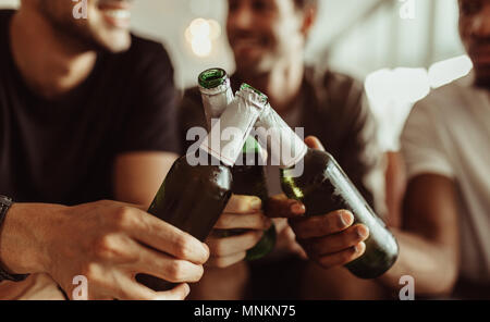 Chiudere fino a tre amici tostare le loro bottiglie di birra mentre avente una parte a casa. Gli uomini elevando le bottiglie per un brindisi seduti a casa. Foto Stock