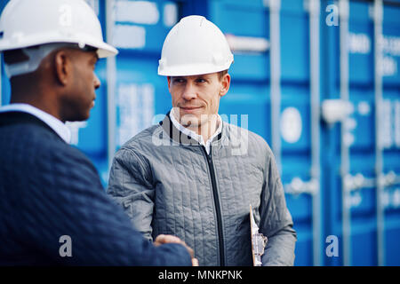 Due ingegneri indossando hardhats agitando mani insieme mentre in piedi da contenitori di spedizione su un dock commerciale Foto Stock