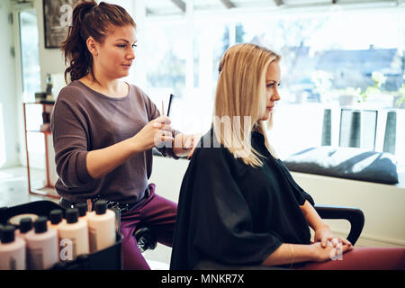 Giovani parrucchiere il taglio dei capelli di una giovane donna cliente seduto in una sedia mentre si lavora nel suo salon Foto Stock