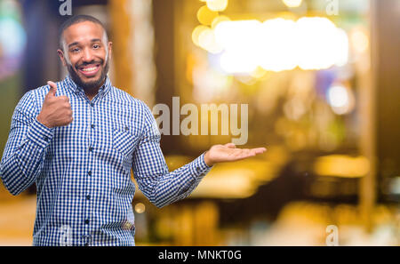 African American uomo con barba pollice in alto tenendo qualcosa in mano vuota e rendendo ok gesto di notte Foto Stock