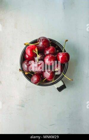 Organico mature appena raccolte le ciliegie dolci in smalto Vintage Mug in metallo grigio pietra sullo sfondo di calcestruzzo dalla finestra. Tavolo da giardino. Lay piatto. Estate Har Foto Stock