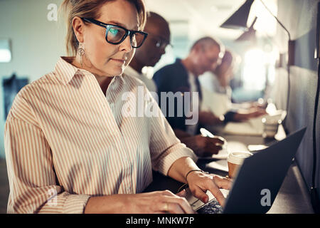 Femmina matura designer che lavora su un computer portatile mentre è seduto in una fila con i colleghi a una tabella in un ufficio moderno Foto Stock