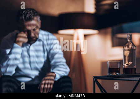 Bottiglia e vetro con alcol in primo piano e sprechi addict seduti nella parte posteriore (sfocata) Foto Stock