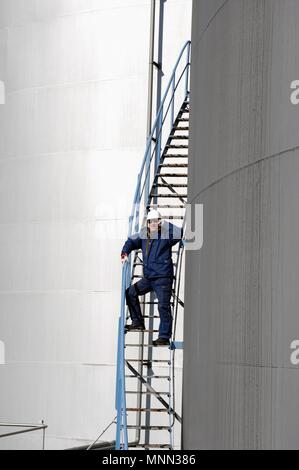 Lavoratore di raffineria in piedi sulla scaletta parlando al telefono accanto a olio industriale e serbatoi di accumulo di carburante. Foto Stock