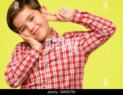 Bel bambino toddler verde con occhi che guardano a telecamera attraverso le dita nel gesto di vittoria con una strizzatina d'occhio l'occhio e soffiando un bacio su sfondo giallo Foto Stock