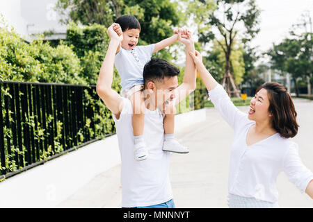 Carino padre asiatici piggybacking suo figlio insieme con sua moglie nel parco. Famiglia eccitato alzando le mani insieme con la felicità Foto Stock