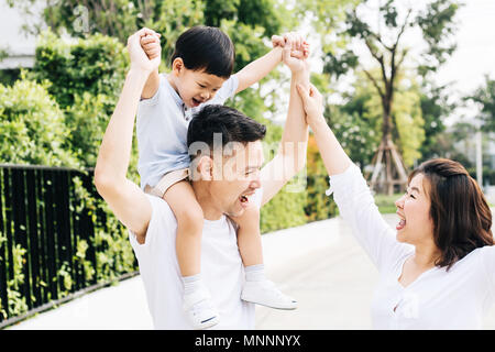 Carino padre asiatici piggybacking suo figlio insieme con sua moglie nel parco. Famiglia eccitato alzando le mani insieme con la felicità Foto Stock