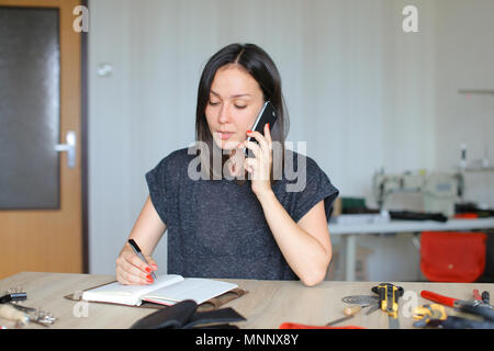 Craftswoman caucasica parlando da smartphone e scrivere a mano in pelle per portatile a casa. Foto Stock