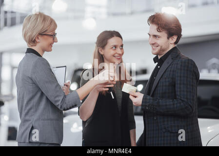 Di acquistare la loro prima vettura insieme. Angolo di Alta Vista del giovane venditore auto in piedi presso la concessionaria dicendo circa le caratteristiche della vettura per i clienti Foto Stock