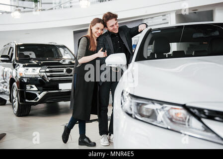 Di acquistare la loro prima vettura insieme. Angolo di Alta Vista del giovane venditore auto in piedi presso la concessionaria dicendo circa le caratteristiche della vettura per i clienti Foto Stock