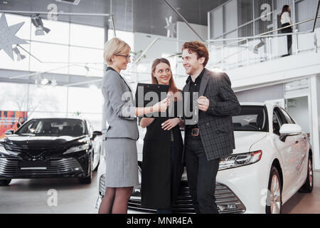 Di acquistare la loro prima vettura insieme. Angolo di Alta Vista del giovane venditore auto in piedi presso la concessionaria dicendo circa le caratteristiche della vettura per i clienti Foto Stock