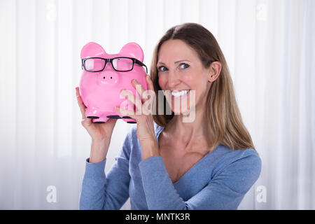 Sorridente donna matura Azienda Piggybank rosa con occhiali neri contro persiane bianche Foto Stock