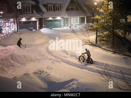 Non è un giorno per un giro in bici: Un ciclista spinge attraverso la neve profonda su una strada di notte. Una donna si cancella un vialetto. Foto Stock