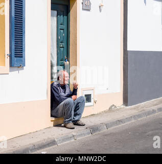 Godendo di un fumo: un uomo si siede sul gradino all'ingresso a casa sua e gode di una sigaretta nel sole mattutino. Foto Stock