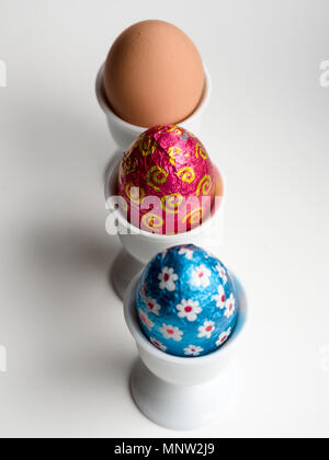 Le uova di Pasqua di cioccolata e un marrone di un uovo di gallina uova in coppe: una fila di tre uova in tre bianchi d'uovo in porcellana tazze su uno sfondo bianco. Foto Stock