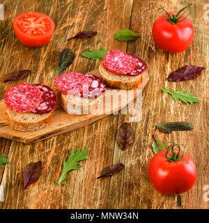 Sandwich fatti in casa con salsiccia affumicata, il salame e i pomodori sul vecchio tavolo in legno. Thinly slice piccante. Tipico italiano o spagnolo snack. Square. Close-up Foto Stock