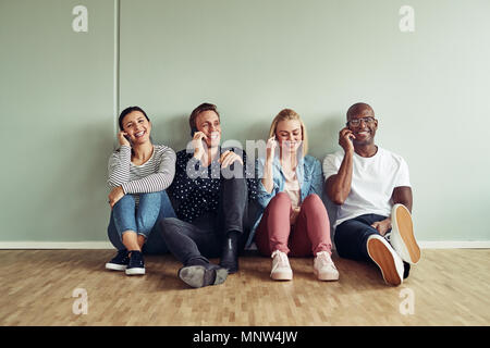 Gruppo sorridente di diversi colleghi seduti insieme su un ufficio piano parlando sui loro telefoni cellulari durante una pausa in un ufficio moderno Foto Stock