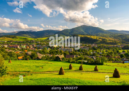 Bellissimo paesaggio rurale in montagna. haystacks sulle colline erbose. villaggio ai piedi della montagna. interessante formazione delle nuvole sopra il bordo Foto Stock