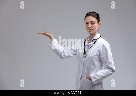 Medico donna di infermiere che indossa uno stetoscopio e camice estendendo la sua mano vuota per la collocazione di prodotti isolati su grigio Foto Stock