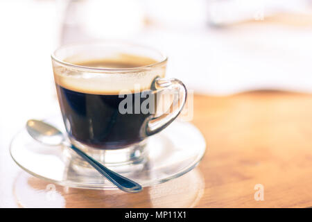 Una tazza calda di caffè nero in vetro chiaro cup con un piccolo cucchiaio d'argento sul tavolo di legno in condizioni di intensa luce mattutina, messa a fuoco selettiva e spazio di copia Foto Stock