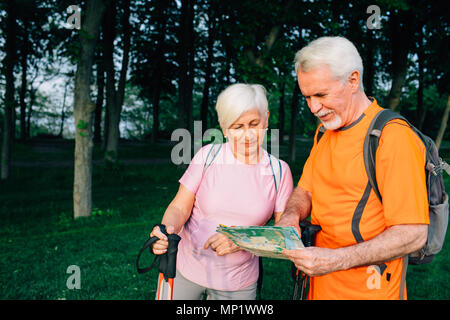 Coppia senior controllo mappa, in piedi con pali trekking in foresta. il pensionamento, di viaggio e di uno stile di vita attivo degli anziani Foto Stock