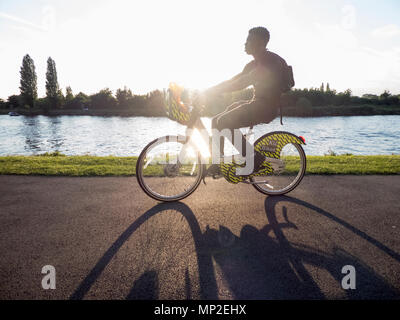 Giovane uomo retroilluminato in bicicletta lungo il Tamigi su Kingston University e-bike con lunga ombra Foto Stock