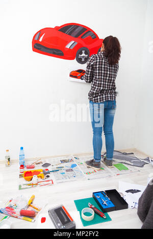 Close-up di una giovane donna artista e la madre del ragazzo per il disegno di un bambino su un muro bianco un bel rosso auto in una luce stanza dei bambini, sul pavimento giace Foto Stock