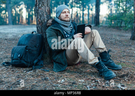 Adulto maschio bello traveler con zaino siede in autunno pineta vicino albero, mantenendo compressa la ricreazione in appoggio di viaggio il concetto di destinazione Foto Stock