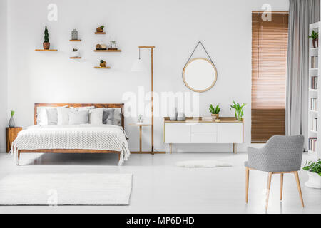 Spaziose bianco designer interiore camera da letto con letto in legno con la biancheria per il letto e i cuscini, night-table, piccoli ripiani sopra e poltrona moderna sulla destra. Foto Stock