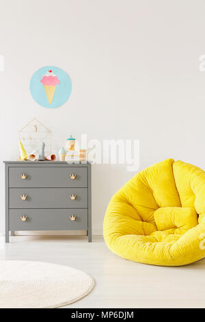 Pouf giallo accanto al grigio di una cassettiera e gelato poster su una parete bianca in kid room interior. Luogo ideale per il tuo poster Foto Stock