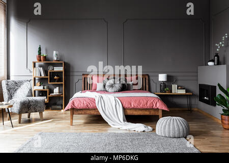 Foto reale di un accogliente interiore camera da letto con letto in legno nel mezzo, camino sulla destra e grigio, poltrona modellata sulla sinistra Foto Stock