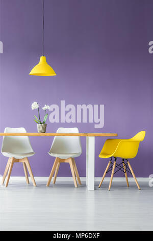 Pavimento bianco e viola la parete soggiorno interno con semplici, colore contrastante di sedie, tavolo con una pianta in un vaso e appeso un lampada gialla Foto Stock