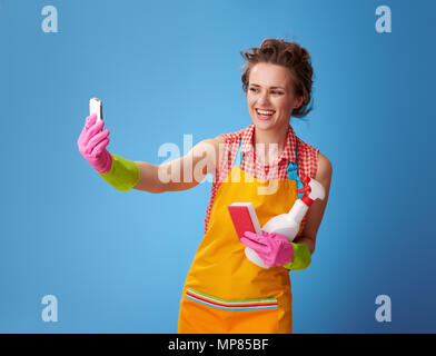 Grande tempo di pulizia. Sorridendo casalinga moderna in un grembiule giallo con cucina in spugna e di una bottiglia di detergente tenendo selfie con lo smartphone contro blu Foto Stock