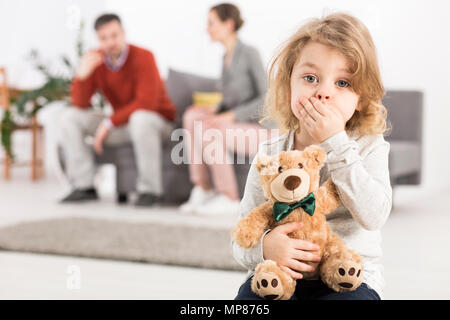 Preoccupato per il ragazzo con Teddy bear, tenendo la mano sulla sua bocca, in background l uomo e la donna seduta sul divano Foto Stock