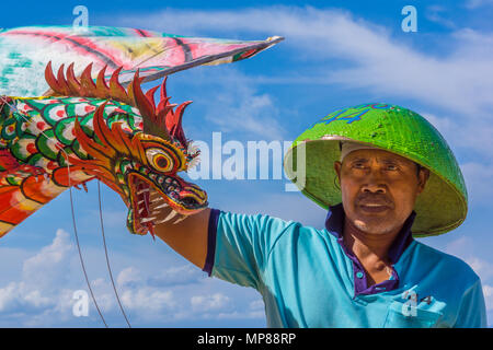 Un uomo indonesiano in verde cappello tenendo un grande aquilone rosso con un terribile drago di testa, Sanur, Bali, Indonesia, 21 aprile 2018 Foto Stock