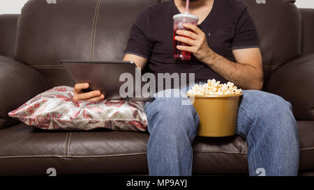 Singolo uomo guarda il filmato sulla tavoletta digitale. Essi sono di bere succo di frutta e mangiare popcorn. Seduti sul divano del soggiorno. Foto Stock