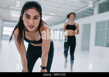 Donna sensazione di stanchezza fisica durante la sessione di allenamento in palestra. Femmina in sportswear flessione in avanti con le mani sulle ginocchia con altre donne che esercitano in b Foto Stock